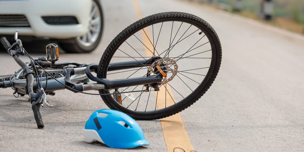 A bicycle accident scene shows a blue helmet and eyeglasses lying on the road next to a white car, hinting at a recent collision.
