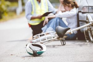 A helmet lies on the road near a bicycle after a bicycle accident, with a woman sitting beside a car and another person in a reflective vest taking notes.