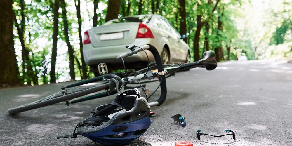 A silver car and a fallen bicycle lie on a tree-lined road after a bicycle accident, with a helmet and broken sunglasses scattered nearby.