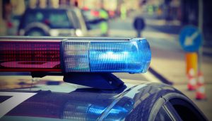 Close-up of a police car's flashing blue and red lights, with a blurred street scene and traffic signs in the background, suggesting emergency response to a nearby bicycle accident.