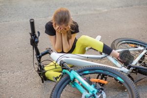 A person in yellow leggings sits on the ground next to a fallen bike, covering their face with their hands after a bicycle accident.