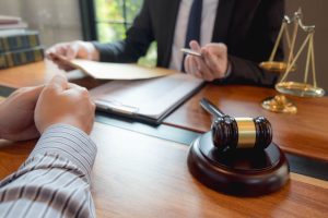 Two people in a professional setting discuss legal documents at a desk with a gavel, balance scale, and paperwork visible.