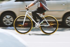 A person rides a white bicycle with yellow rims on a city street, while a beige car passes by in the background—capturing a moment where staying alert can help prevent a potential bicycle accident.