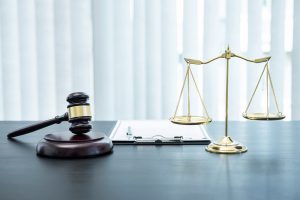 A wooden judge’s gavel, a gold balance scale, and a clipboard sit on a dark desk in front of vertical blinds—an atmosphere well-suited for discussing a hit-and-run bicycle accident case.