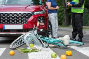  A fallen bicycle and scattered groceries lie on a crosswalk in front of a red car, while two people stand nearby—one in a reflective vest holding a notepad, possibly documenting the scene for Common Causes of Bicycle Accidents.