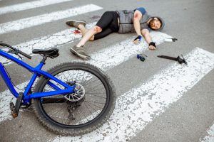  A cyclist wearing a helmet lies injured on a crosswalk after a hit-and-run bicycle accident, with a blue bicycle and personal items scattered nearby.