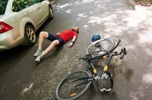 A man lies on the road near a bicycle and a car, with a helmet beside him, suggesting a recent bicycle accident or collision.