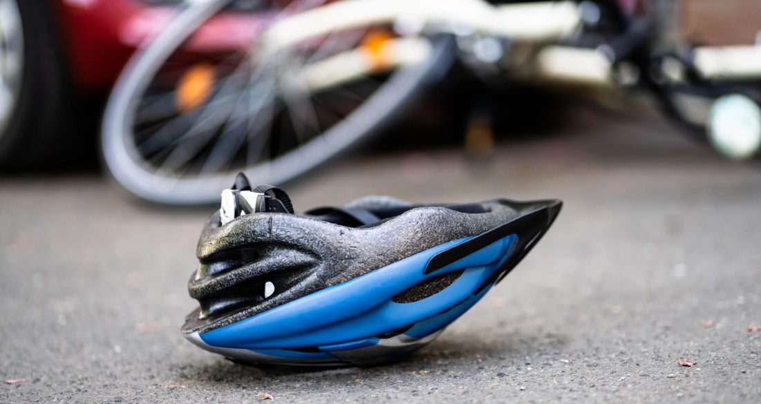A blue bicycle helmet lies on the pavement, suggesting a recent bicycle accident, with a fallen bicycle and a red car in the background.