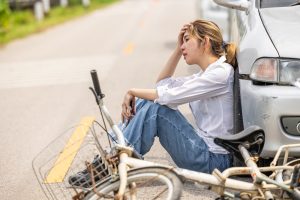A woman sits on the road leaning against a car, looking distressed, next to a damaged bicycle after a bicycle accident.