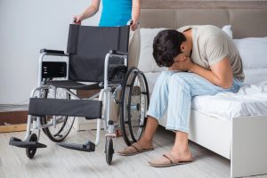  A man sits on a bed with his head in his hands, looking distressed after a motorcycle accident, while another person stands behind an empty wheelchair nearby.
