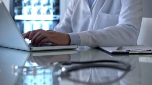 A person in a white lab coat types on a laptop at a desk with a clipboard, stethoscope, and X-ray images from a recent motorcycle accident in the background.