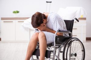 A person in a hospital gown sits in a wheelchair, leaning forward with their face buried in their hands after a motorcycle accident in a clinical room.