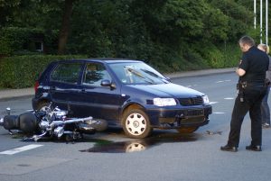 A police officer stands near a blue car and a fallen motorcycle after an apparent motorcycle accident on a street, with liquid spilled on the road.