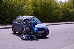 A blue car and a blue motorcycle have collided in a motorcycle accident at an intersection, with the motorcycle lying on its side in front of the car. Trees and pavement are visible in the background.