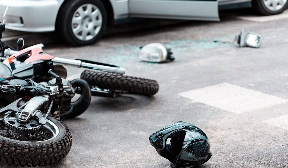 A motorcycle accident is evident as a motorcycle lies on its side near a helmet and broken glass beside a car with an open door, indicating a recent collision.