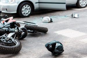 A motorcycle accident is evident as a motorcycle lies on its side near a helmet and broken glass beside a car with an open door, indicating a recent collision.