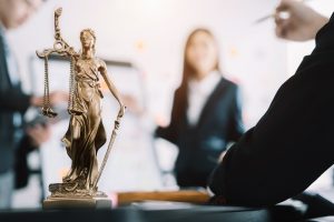 Bronze Lady Justice statue on a desk, with three blurred people in business attire in the background discussing a motorcycle accident.