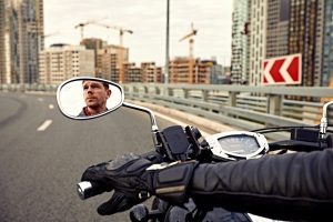 A person rides a motorcycle on a city road, visible in the rearview mirror, with construction cranes and tall buildings in the background, highlighting the potential risks of a motorcycle accident in busy urban environments.